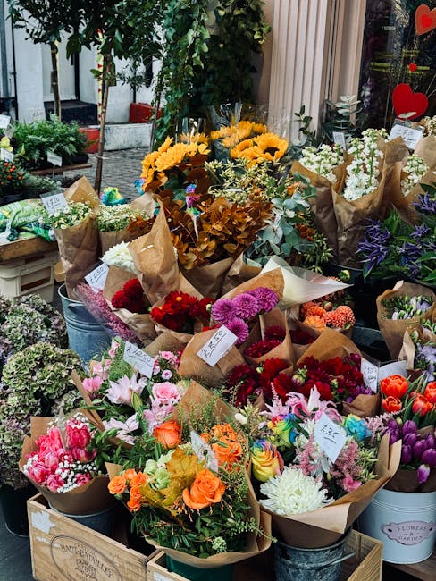 An outdoor display of a variety of fresh floral bouquets arranged on a wooden crate and metal buckets in front of a shop window. The bouquets feature vibrant flowers including pink lilies, orange rose
