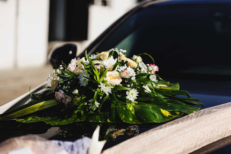 A floral arrangement featuring a bouquet of fresh white roses, daisies, and small pink blossoms, accented with lush green foliage and long leaves. The bouquet is placed on the hood of a black car, par