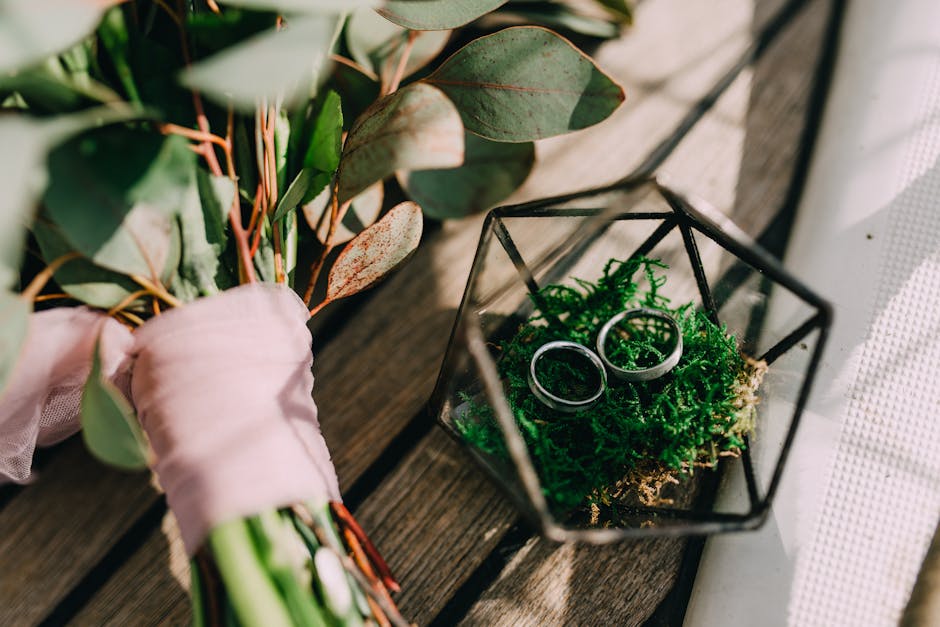 A close-up of a bouquet of fresh greenery and eucalyptus leaves, wrapped in soft pink ribbon, resting on a wooden surface. Next to the bouquet is a geometric glass terrarium containing bright green mo