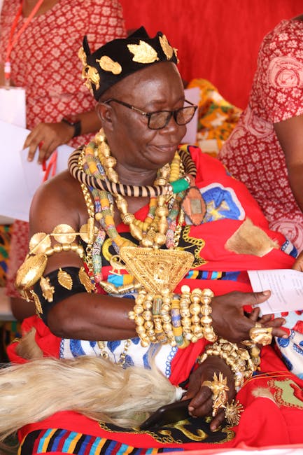 A woman dressed in traditional African attire decorated with numerous gold jewelry pieces, including necklaces, bangles, and earrings, sits indoors against a vibrant red backdrop. She wears a black he