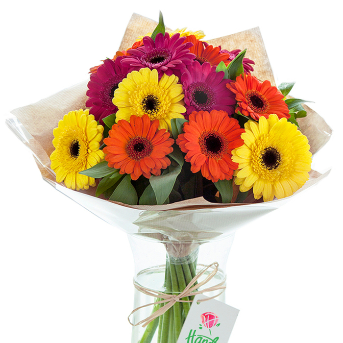 In a scene awash with brightness and joy, a hand-tied bouquet of Gerbera daisies commands attention-each bloom hand-selected by a Tulse Hill florist with a keen eye for balance and beauty. The striking colorful array brings together intense sunny yellows, passionate oranges, and deep fuchsia tones, with petals radiating outward from rich, velvety dark centers. Every stem is thoughtfully nestled in opulent greenery, providing an intricate backdrop suggestive of the lush gardens around Brockwell Park. Wrapped in a layer of natural kraft paper with soft, visible texture, then finished with an overlay of clear, gently crinkled cellophane, the bouquet achieves both rustic elegance and protective luxury. The clear glass vase beneath holds the stems upright, water glinting through its clean lines, ensuring the flowers' enduring vitality. A simple piece of earthy twine encircles the stems at the neck, tied in a careful knot supporting a delicately suspended white tag graced with a red rose motif-hinting at the arrangement's special intent. Designed for brightening anniversaries, birthdays, or act as a centrepiece for village gatherings in Tulse Hill, this arrangement encapsulates the professional skill and emotion of a seasoned local florist. The interplay of colour, form, and texture in this arrangement evokes both celebration and gratitude-a floral gift that brings the heart of Tulse Hill into any home.