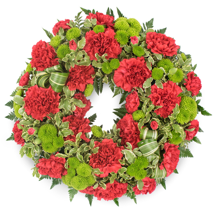A richly detailed funeral wreath fills the frame, its perfectly round shape set crisply against a clean white background that highlights every petal, leaf, and ribbon. The wreath is an intricate blend of deep red and glowing lime green, with the flowers arranged so closely together that the circle feels almost like a living tapestry. Red carnations form the main body of the design; their petals are densely frilled and soft-looking, creating layers of velvety texture all the way around the ring. Some blooms are fully open, showing ruffles within ruffles, while others are in earlier stages of opening, their edges gently curving inward, which adds a natural variation and sense of depth. Between the carnations sit clusters of lime green button chrysanthemums, small and perfectly rounded, like bright beads of colour. Each button mum is made up of countless tiny petals pressed closely together, giving the wreath a playful yet refined contrast in both texture and tone. The foliage foundation is lush and carefully considered. Dark green leatherleaf fern extends just beyond the flowers, forming a feathered border that frames the wreath. Interwoven are sprigs of variegated pittosporum, with dark centres and creamy margins that lighten the overall effect and soften transitions between blooms. A striped green ribbon, fashioned into gentle loops near the base, echoes both the light and dark greens of the foliage, subtly enhancing the design. The neutral background and soft, even lighting create a tranquil mood, allowing this classic tribute to be imagined resting in a quiet Tulse Hill chapel, at a crematorium service, or on a graveside, offering a dignified expression of sympathy and remembrance.