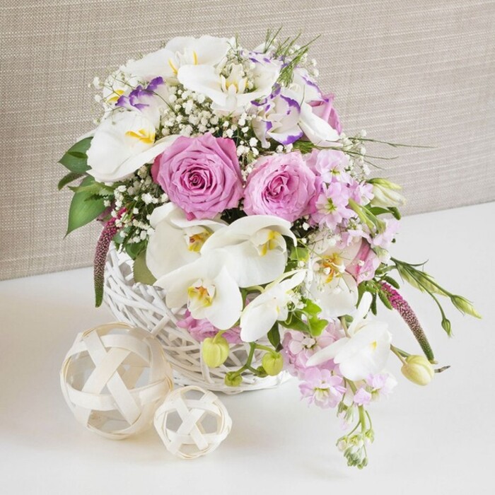 In this serene wedding-inspired image, a low, round white wicker basket is completely filled with an abundant mix of pink and white flowers, creating a lush focal point that would sit perfectly on a Tulse Hill reception table. Large, pastel pink roses open generously across the top of the arrangement, their layered petals spiralling inward and catching soft ambient light, lending a classic and romantic feel. Threaded through the roses are elegant white Phalaenopsis orchids, their broad petals almost translucent at the edges, with warm yellow centers touched by slender strokes of soft purple, adding a refined and slightly exotic note. Interspersed among these statement blooms are delicate white Lisianthus, some with edges brushed in lavender, their ruffled petals giving the composition a gentle movement, as if stirred by a breeze drifting up from nearby Hillside Gardens. Stems of pale pink stock cascade gently over the sides of the basket, introducing vertical rhythm and a subtle, sweet fragrance one can easily imagine. Wispy clouds of white Gypsophila lace between all the larger flowers, lightening the overall effect and giving the bouquet a dreamy, cloud-like softness. Fresh greenery is tucked carefully throughout, with small leaves and tender buds adding contrast, structure, and a sense of natural growth. The wicker basket stands on a spotless white surface, likely a wedding or anniversary table, with two decorative woven spheres in pale natural wood placed close by. Their intricate, interlocking strands echo the basket's texture and introduce an additional tactile element. Behind it all, a warm beige textile backdrop, reminiscent of fine linen, keeps the scene calm and neutral, allowing the floral colours and craftsmanship to truly shine, ideal for couples planning elegant celebrations around Tulse Hill and neighbouring Herne Hill.