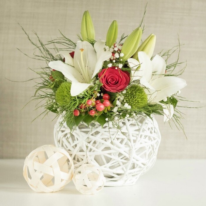 On a bright white tabletop, a refined floral centerpiece is arranged with the quiet confidence of an experienced Tulse Hill florist preparing a wedding table design. The composition begins with a round white vase, crafted from crisscrossing open-weave strands that resemble sculpted wicker or delicately intertwined branches, its airy pattern adding interest even before the flowers are seen. Rising generously from this vessel, several white lilies take centre stage, their large, graceful petals opening outwards to reveal subtle freckles and prominent yellow stamens, suggesting both purity and gentle fragrance. Some lilies remain in slender green bud, their tips just beginning to part, promising that the arrangement will continue to evolve over the coming days. Threaded through the lilies are rich, velvety red roses; one especially striking bloom sits near the middle with petals tightly curled into a near-perfect spiral, embodying classic romance in deep crimson. Accents of bright green pompon-style blooms-likely green chrysanthemums or dianthus-form rounded, cushion-like tufts that pop vividly against the white petals and dark foliage. Tiny sprays of airy white baby's breath permeate the arrangement, creating a soft, cloud-like haze, while small clusters of red berries introduce a festive sparkle that would complement a winter wedding reception near West Norwood or Streatham Hill. A mixture of wispy, fern-like fronds and darker broad leaves spills naturally beyond the edge of the vase, adding fullness and a slightly wild, garden-fresh character. To the left, two pale woven spheres in natural wood tones rest on the surface, echoing the vase's texture and reinforcing the organic theme. Behind it all, a softly textured neutral wall provides a calm backdrop, letting the interplay of white, red, and green speak of celebration, love, and refined style for special gatherings across Tulse Hill.