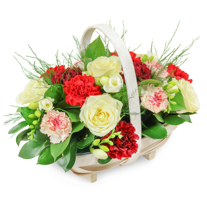A classic condolence flower basket is shown in crisp detail, placed against a bright white background so that every colour and curve can be clearly imagined. The basket itself is crafted from light, natural wood with a shallow oval profile, slender vertical slats, and four small feet that lift it delicately off the surface. An elegant arched handle rises above, painted or finished in a soft off-white tone that feels calm and unobtrusive. Overflowing from this base is a rounded, dome-shaped mass of flowers, so generously filled that the blooms seem to spill just over the edges, creating a sense of abundance and care. Dominating the arrangement are several creamy white roses, fully unfurled, their layered petals glowing gently, symbolising peace, remembrance, and a quiet farewell. Interspersed among them are deep red carnations with densely ruffled petals, their rich colour adding a heartfelt intensity that speaks of enduring love and respect. Softer notes appear in the form of variegated or pink-tipped carnations, where pale pink and white petals are dusted or edged with red, easing the contrast between the pure whites and the stronger crimson tones. Tucked between the larger flowers are clusters of small white blooms, likely freesias, some in bud and some open, their slender stems arching gracefully and suggesting a light, fresh fragrance. The greenery is abundant but carefully placed: broad, glossy leaves frame the composition, while fine, feathery fern-like sprigs push through the top and sides, giving the tribute a slightly untamed, natural softness. The overall effect is warm and dignified, ideally suited to a funeral, memorial, or to be sent as a sympathy gift to a family home in Tulse Hill, perhaps along one of the quiet streets leading towards West Norwood, where a touch of floral comfort can make a difficult day feel a little less stark.