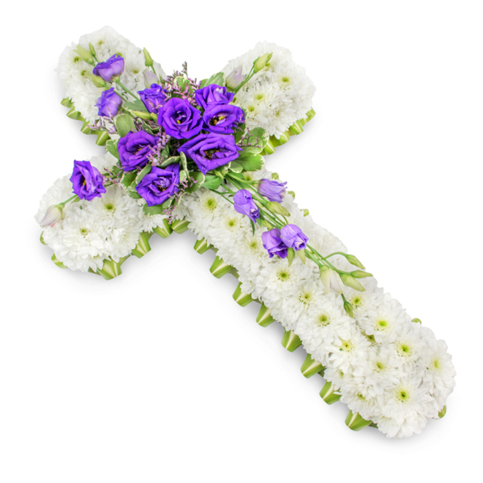 A traditional funeral flower cross, captured from a high, slightly angled viewpoint, rests against a spotless white background that highlights every nuance of the design. The entire cross is carpeted in tightly packed white chrysanthemums, each small flower carefully positioned to eliminate any gaps, creating a smooth, continuous field of white that feels soft and cloud-like. Their tiny green centres form a subtle pattern that breaks up the surface just enough to keep the eye moving. Along the outer perimeter of the cross, a slim lime-green ribbon runs in an unbroken line, hugging every curve and corner. This delicate border adds a fresh, spring-like note and defines the shape crisply, almost as if it were traced in light. At the heart of the cross, where families might focus during a farewell service in Tulse Hill or nearby West Norwood, a rich cluster of deep purple lisianthus blossoms provides a strong visual and emotional anchor. The lisianthus heads range from fully open to gently unfurling, their petals forming layered spirals that look velvety and almost luminous against the white chrysanthemum base. Sprigs of soft green foliage and tiny accent flowers are interwoven among the purple blooms, adding depth, contrast, and a sense of natural movement. From this central spray, additional lisianthus stems drift diagonally down the lower-right arm, some blossoms open, others in bud, echoing the idea of a life's journey continuing beyond the moment of parting. A mild, diffused shadow under the arrangement lifts it ever so slightly from the background. The overall effect is calm, reverent, and deeply respectful, a floral cross that speaks of faith, remembrance, and love held close.