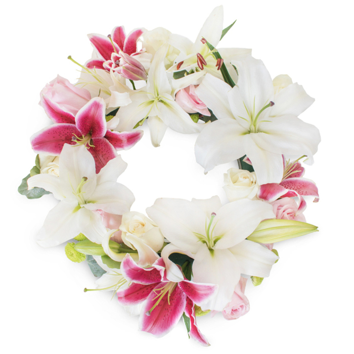A luminous floral wreath, thoughtfully arranged by skilled Tulse Hill florists, takes center stage against a pure white backdrop-its full circle brimming with serene white lilies and radiant fuchsia pink blooms. The pristine, star-shaped lilies unfold gracefully, each petal curving outward to reveal delicate green and yellow stamens, offering an air of tranquility and purity. Splashes of bold fuchsia-pink Stargazer lilies are artfully scattered among the white, creating visual harmony and recalling the lively color seen around Norwood Road's community gardens. These vivid lilies, peppered with subtle speckles toward their centers, introduce depth and drama, guiding the viewer's eye across the entwined ring. Gentle blush pink and creamy white roses cluster between the lilies, some petals tightly hugged in bud form, others softly ruffed in full bloom, their texture and softness diffusing the arrangement's visual impact like a gentle embrace. Glimpses of soft green foliage and hints of eucalyptus peek through the blooms, providing supporting structure and a naturally fresh ambiance. The wreath's composition, a continuous circle, embodies unity, memory, and the cyclical nature of life-qualities perfectly suited for sympathy, funeral tributes, or celebration of life services served throughout Tulse Hill. Overlaying the careful artistry is a profound reverence, manifesting care and compassion for loved ones, as well as the quiet resilience found within the community.