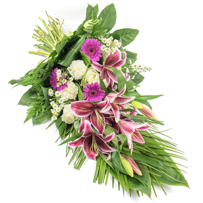 This elongated funeral sheaf is displayed against a clean white background, its diagonal line running from the gathered stems at the upper left to a gentle cascade of leaves and blooms at the lower right, creating a soft crescent that evokes a tear of remembrance. At the heart of the design, several magnificent Stargazer lilies unfurl, their petals saturated in vivid magenta-pink with crisp white margins and dark burgundy speckles radiating from the centre. Their prominent stamens and lightly curved petals give the flowers a sense of movement, as if they are still opening. Some lilies remain in bud form, green-tipped and blushed with pink, reminiscent of memories still forming in the minds of those left behind. Tucked close to them are creamy white roses, some tight and conical, others beginning to reveal their layered hearts, expressing reverence, love, and farewell. Ruffled white carnations add a soft, cushion-like texture and underscore the feeling of enduring affection. Bright magenta gerbera daisies introduce a modern splash of colour, offering a touch of hope and warmth amid the more classic blooms. Threaded between them, small clusters of delicate white daisy-style fillers lighten the overall look and draw the eye along the length of the arrangement. Surrounding the flowers, a rich collar of greenery brings structure and depth: broad, glossy leaves provide a firm base, while airy fern fronds and long, arching blades of green foliage emphasize the flowing, tapered shape. The lighting is bright and even, highlighting every petal and leaf without casting harsh shadows. Crafted with the sensitivity of a local Tulse Hill florist who understands the needs of families in the area, this substantial sympathy spray is perfect for a funeral, chapel service, or memorial gathering, echoing both the tenderness and strength found in the community around nearby Herne Hill and West Norwood.