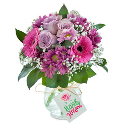 Step into a moment of heartfelt storytelling with this vivid Tulse Hill bouquet-an artful expression of affection, joy, and the comfort of familiar places. The focal point is a cluster of pale lilac roses, their gently curled petals evoking the soft dawn light streaming across leafy neighbourhood gardens. Circling these, vibrant magenta Gerbera daisies stand tall, their bold, open blooms framed by dark-eyed centres that invite lingering glances, reminiscent of the lively stalls at Herne Hill Market. Smaller plum-coloured chrysanthemums add layers of texture and subtle wildness, while baby's breath scatters tiny white blossoms throughout, like dappled sunlight dancing over Dulwich Wood. Glossy, verdant green leaves anchor the bouquet, giving a sense of movement and depth. The arrangement is nestled in a clear, textured glass vase with a unique 'cracked ice' effect-its artisanal detail reflecting the creative spirit of local Tulse Hill florists. A raffia-tied tag provides a personal touch, ready to bear words of celebration or sympathy alike. This composition feels both modern and intimate, especially suited to marking a significant milestone or simply letting someone in SE27 know they are cherished, with every petal whispering the dedication and artistry of Tulse Hill's floral community.