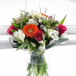 This luxurious bouquet, expertly crafted in Tulse Hill, starts with a radiant orange Gerbera at its heart-its vivacious hue radiates warmth, recalling the lively buzz of the high street on market day. Surrounding it, deep magenta and crimson roses offer a timeless richness; the open blooms and newly awakening buds create a visual symphony of romance and anticipation, like the changing seasons glimpsed in Brockwell Park. Lacy clusters of white chrysanthemums and delicately striped alstroemeria add layers of crisp contrast and volume, capturing the uplifting feeling of early spring in a window-lit Tulse Hill flat. Towards the top, creamy pale roses and subtle lavender florets lend softness and an elusive, comforting scent. The intricate arrangement stands in a clear glass vase, water sparkling around the green stems, as bands of glossy Monstera and slender, spiky leaves frame the flowers, echoing the leafy charm of village greens and residential gardens in SE24. The result is an exuberant, fresh bouquet-an ideal gift for celebrations, anniversaries or just to lift the everyday with the essence of Tulse Hill's gentle energy and blooming local character.